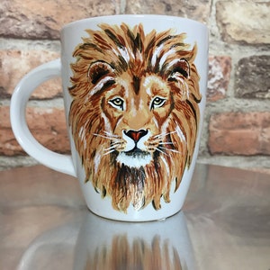 May include: White ceramic mug with a hand-painted image of a lion's face. The lion has a brown mane, a white beard, and a black nose.