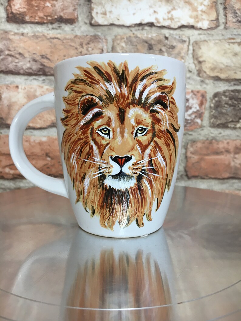 May include: White ceramic mug with a hand-painted image of a lion's face. The lion has a brown mane, a white beard, and a black nose.