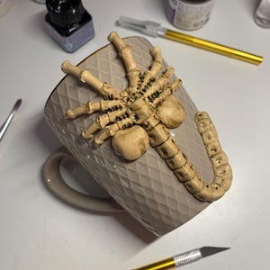 May include: A beige ceramic mug with a diamond pattern, adorned with a detailed, light brown alien facehugger sculpture. The mug has a matching handle and is surrounded by art supplies.