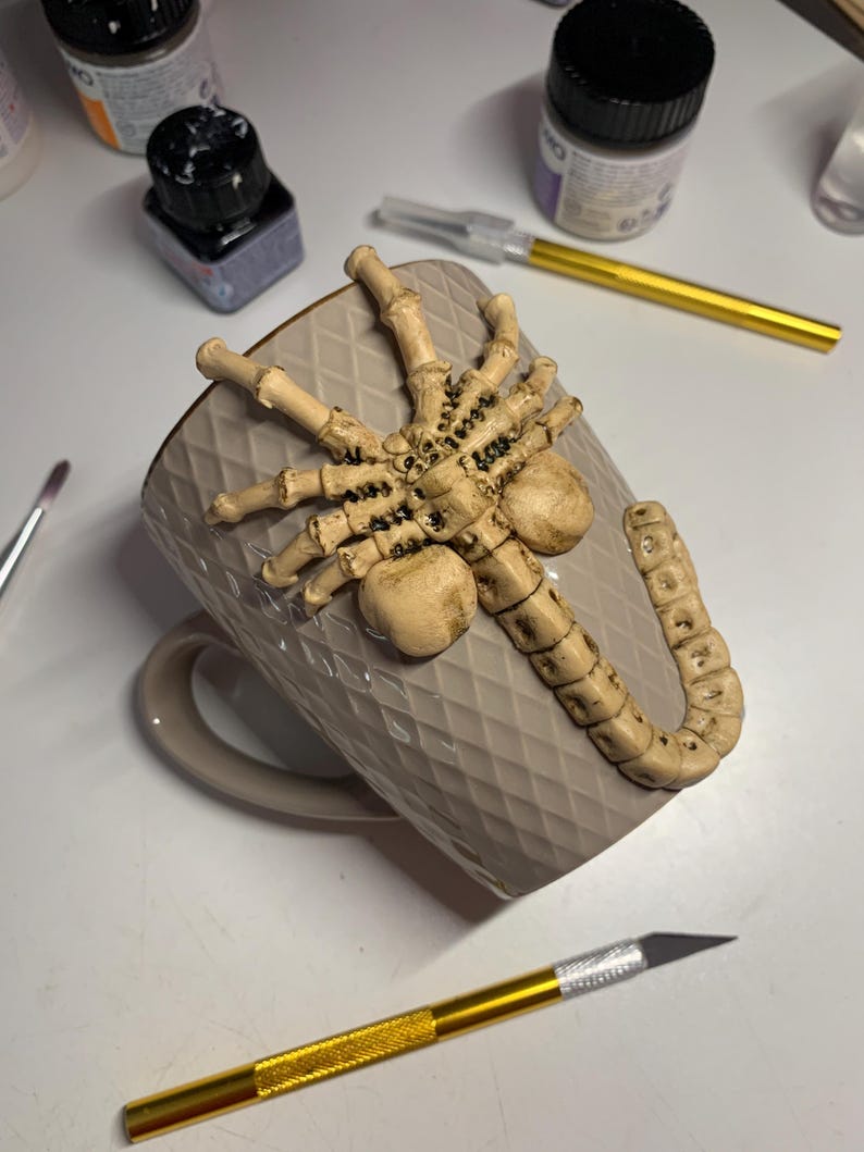 May include: A beige ceramic mug with a diamond pattern, adorned with a detailed, light brown alien facehugger sculpture. The mug has a matching handle and is surrounded by art supplies.
