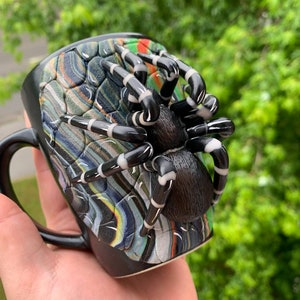 Puede incluir: Taza de cer&aacute;mica negra con un dise&ntilde;o de ara&ntilde;a en 3D. La ara&ntilde;a tiene un cuerpo y patas negras con rayas blancas. El exterior de la taza tiene un patr&oacute;n colorido y texturizado. La taza tiene un asa negra.