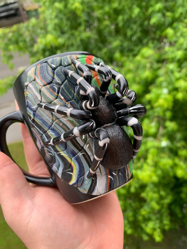 Puede incluir: Taza de cer&aacute;mica negra con un dise&ntilde;o de ara&ntilde;a en 3D. La ara&ntilde;a tiene un cuerpo y patas negras con rayas blancas. El exterior de la taza tiene un patr&oacute;n colorido y texturizado. La taza tiene un asa negra.