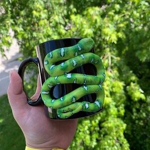 Puede incluir: Taza de cer&aacute;mica negra con una serpiente verde y blanca enrollada. La serpiente tiene una apariencia texturizada y est&aacute; enrollada alrededor de la taza. La taza tiene un asa negra.