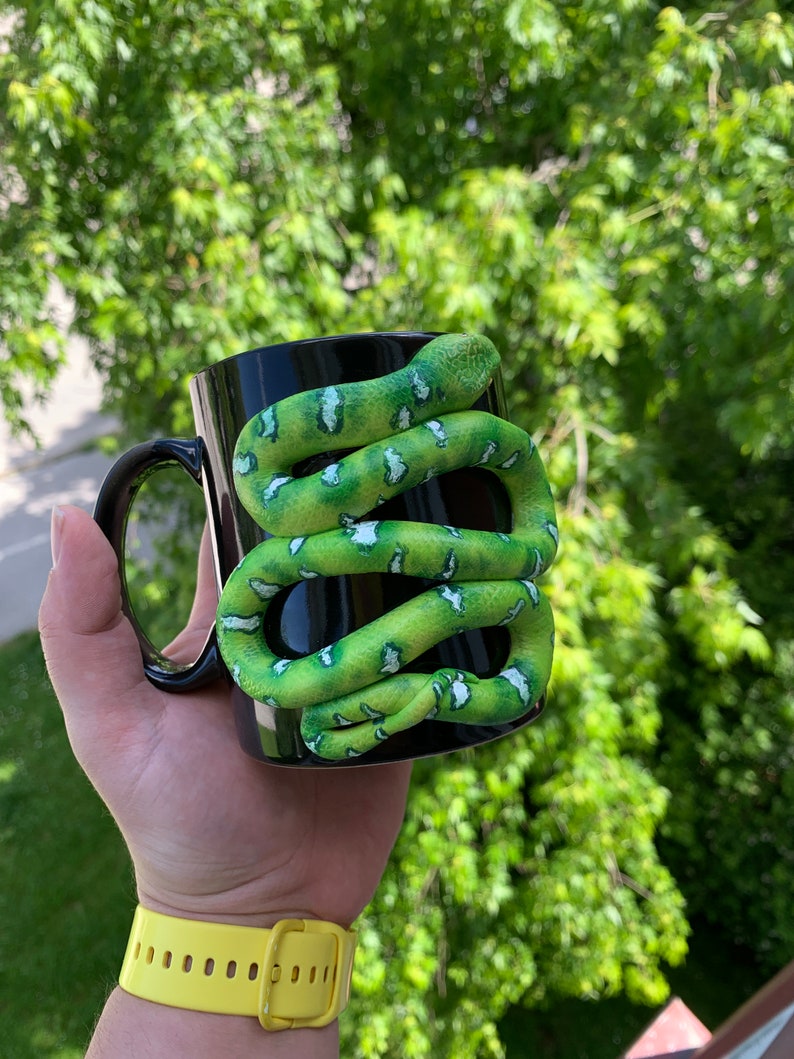 Puede incluir: Taza de cer&aacute;mica negra con una serpiente verde y blanca enrollada. La serpiente tiene una apariencia texturizada y est&aacute; enrollada alrededor de la taza. La taza tiene un asa negra.