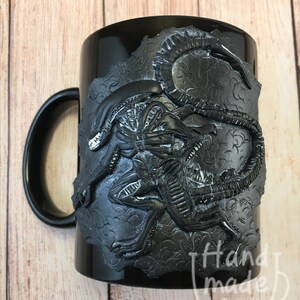 Peut inclure: Mug en c&eacute;ramique noire avec un motif d'extraterrestre sculpt&eacute; en 3D. L'extraterrestre est noir et gris et a une surface textur&eacute;e. La tasse a une poign&eacute;e noire et les mots "Handmade for" sont visibles au bas de la tasse.