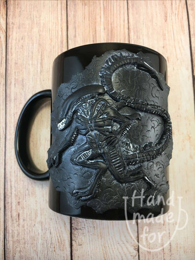 Peut inclure: Mug en c&eacute;ramique noire avec un motif d'extraterrestre sculpt&eacute; en 3D. L'extraterrestre est noir et gris et a une surface textur&eacute;e. La tasse a une poign&eacute;e noire et les mots "Handmade for" sont visibles au bas de la tasse.