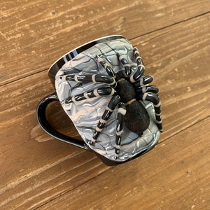 Puede incluir: Taza de caf&eacute; negra con un dise&ntilde;o de ara&ntilde;a en 3D. La taza presenta un asa y un borde negros, con un patr&oacute;n exterior de telara&ntilde;a gris y blanco. Una ara&ntilde;a detallada en blanco y negro est&aacute; adherida al lateral de la taza.
