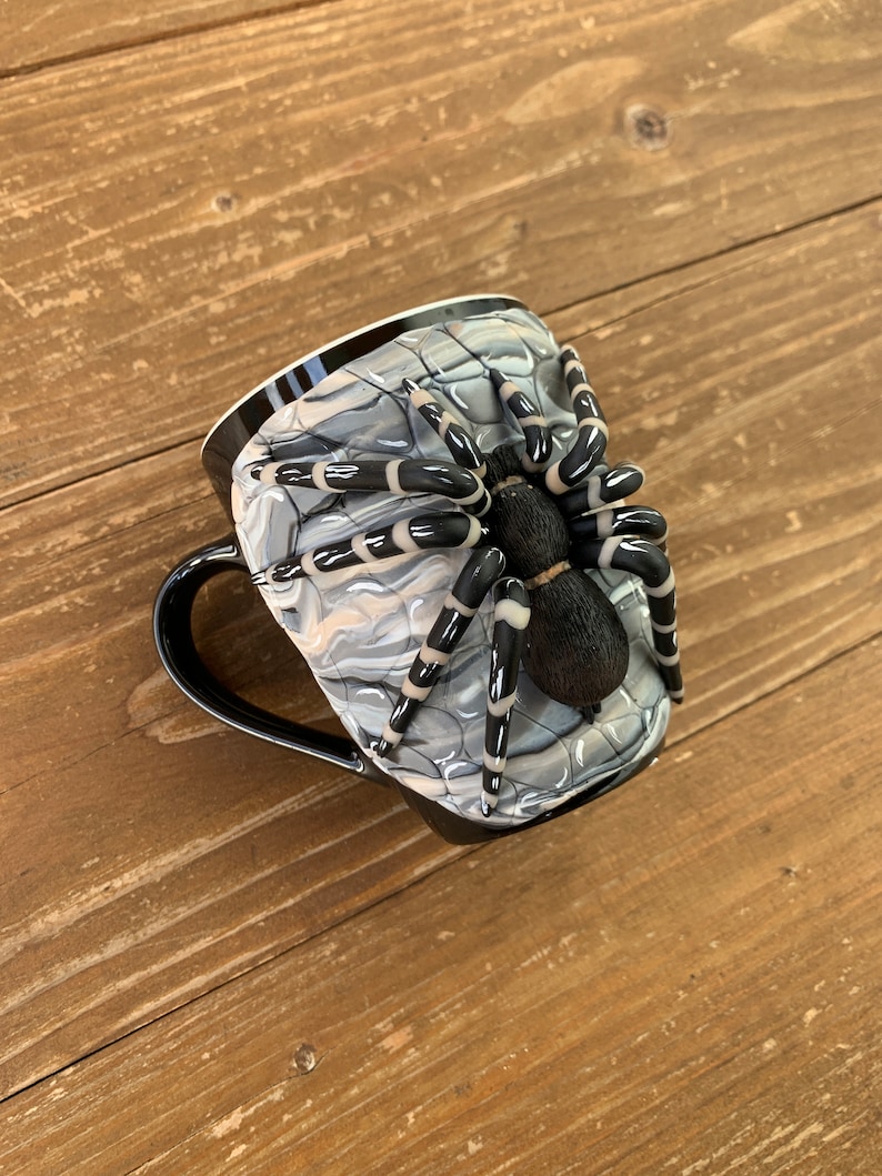 Puede incluir: Taza de caf&eacute; negra con un dise&ntilde;o de ara&ntilde;a en 3D. La taza presenta un asa y un borde negros, con un patr&oacute;n exterior de telara&ntilde;a gris y blanco. Una ara&ntilde;a detallada en blanco y negro est&aacute; adherida al lateral de la taza.