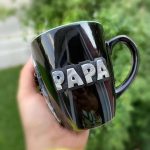 Puede incluir: Taza de cer&aacute;mica negra con la palabra "PAPA" en letras plateadas. La taza tiene un acabado brillante y un asa curvada. La taza se sostiene en una mano, con un fondo verde borroso.