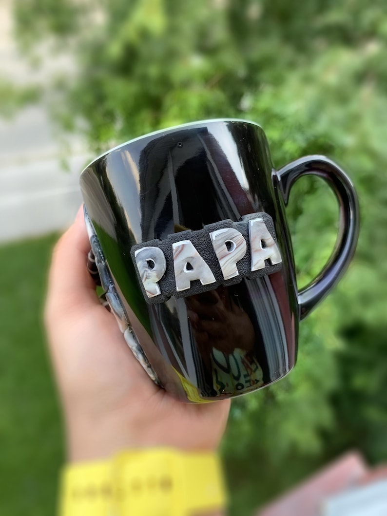 Puede incluir: Taza de cer&aacute;mica negra con la palabra "PAPA" en letras plateadas. La taza tiene un acabado brillante y un asa curvada. La taza se sostiene en una mano, con un fondo verde borroso.