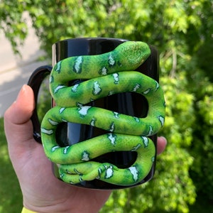 Puede incluir: Taza de cer&aacute;mica negra con una serpiente verde y blanca enrollada alrededor. La serpiente tiene un patr&oacute;n texturizado y est&aacute; enrollada alrededor de la taza. La taza se sostiene en una mano con un reloj amarillo.