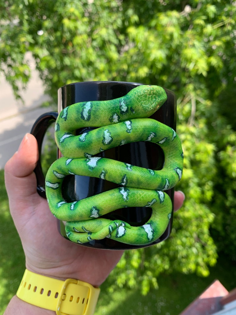 Puede incluir: Taza de cer&aacute;mica negra con una serpiente verde y blanca enrollada alrededor. La serpiente tiene un patr&oacute;n texturizado y est&aacute; enrollada alrededor de la taza. La taza se sostiene en una mano con un reloj amarillo.