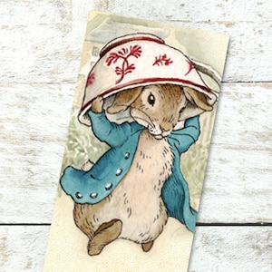 May include: A bookmark featuring a cartoon rabbit wearing a blue jacket and holding a red and white teacup over its head. The bookmark has a quote from the book "The Tale of Peter Rabbit" that reads: "But Peter, who was very naughty, ran straight away to Mr. McGregor's garden, and squeezed under the gate!"