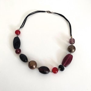 May include: A necklace with a black cord, adorned with a mix of beads in various shapes and colours. The beads are in shades of red, black, brown, and purple. The design features a variety of bead sizes, creating a unique look.