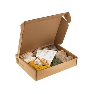 Puede incluir: Una caja de cartón con la tapa abierta, que revela un conjunto de cordones de macramé de colores e instrucciones. Los cordones son en tonos beige, verde y amarillo.