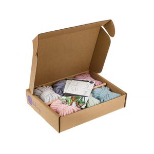 Puede incluir: Una caja de cartón con la tapa abierta, que revela cinco ovillos de cuerda de algodón de colores. La cuerda es en tonos de rosa, azul y gris. La caja está etiquetada con "Macramé" e incluye instrucciones para hacer proyectos de macramé.