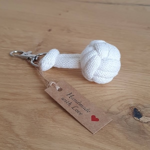 Puede incluir: Llavero de cuerda de algodón blanco con una bola anudada en el extremo. El llavero tiene un cierre de metal plateado y una etiqueta de papel marrón que dice "Handmade with Love" con un corazón rojo.