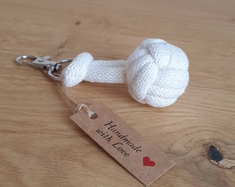 Könnte beinhalten: Schlüsselanhänger aus weißem Baumwollseil mit einer geknüpften Kugel am Ende. Der Schlüsselanhänger hat einen silbernen Metallverschluss und ein braunes Papieretikett mit der Aufschrift "Handmade with Love" und einem roten Herzen.