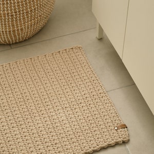 May include: A beige crocheted bath mat with a brown leather strap. The mat is on a tiled floor in a bathroom.
