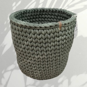 Puede incluir: Una cesta grande de crochet verde con un asa de cuero marrón. La cesta está hecha de hilo grueso y tiene un patrón tejido texturizado.