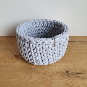 May include: A gray crocheted storage basket with a leather handle. The basket is made of thick cotton rope and has a round shape.