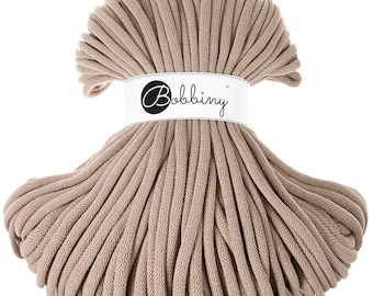 May include: A close-up of a large spool of beige cotton cord. The cord is tightly wound and has a smooth texture. The spool is labeled "Bobbiny".
