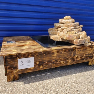 Könnte beinhalten: Ein Holztisch mit einem schwarzen Wasserbecken darauf. Das Wasserbecken besteht aus übereinander gestapelten Steinen und hat einen Wasserfall. Der Tisch ist dunkelbraun gebeizt und trägt ein Schild mit der Aufschrift "Cascade d'Eau Electrique Miniature".