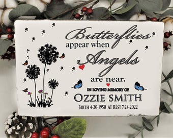 May include: A rectangular white memorial plaque featuring black text that reads Butterflies appear when Angels are near. IN LOVING MEMORY OF OZZIE SMITH BIRTH 4-20-1950 AT REST 7-24-2022. Includes black dandelion illustrations, small blue butterflies, and red hearts.