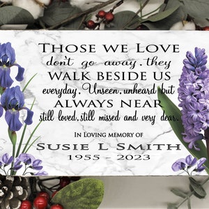 May include: A memorial plaque with a marble-like background, featuring the text "Those We Love" and floral accents. The plaque includes the name "Susie L Smith" and dates 1955-2023, surrounded by purple iris and hyacinth flowers.