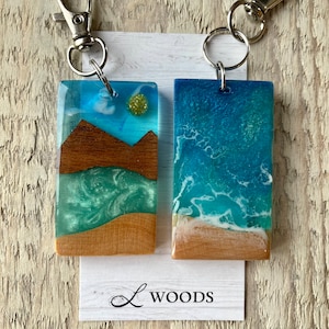 May include: Two resin keychains with a wood frame. One keychain features a blue and green ocean scene with a white sandy beach. The other keychain features a blue sky with a brown mountain scene. Both keychains have glitter embedded in the resin. The keychains are attached to silver key rings.