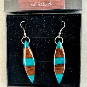 May include: A pair of teal and brown wood earrings with silver hooks. The earrings are in a black box with the text "L Woods" on the lid.