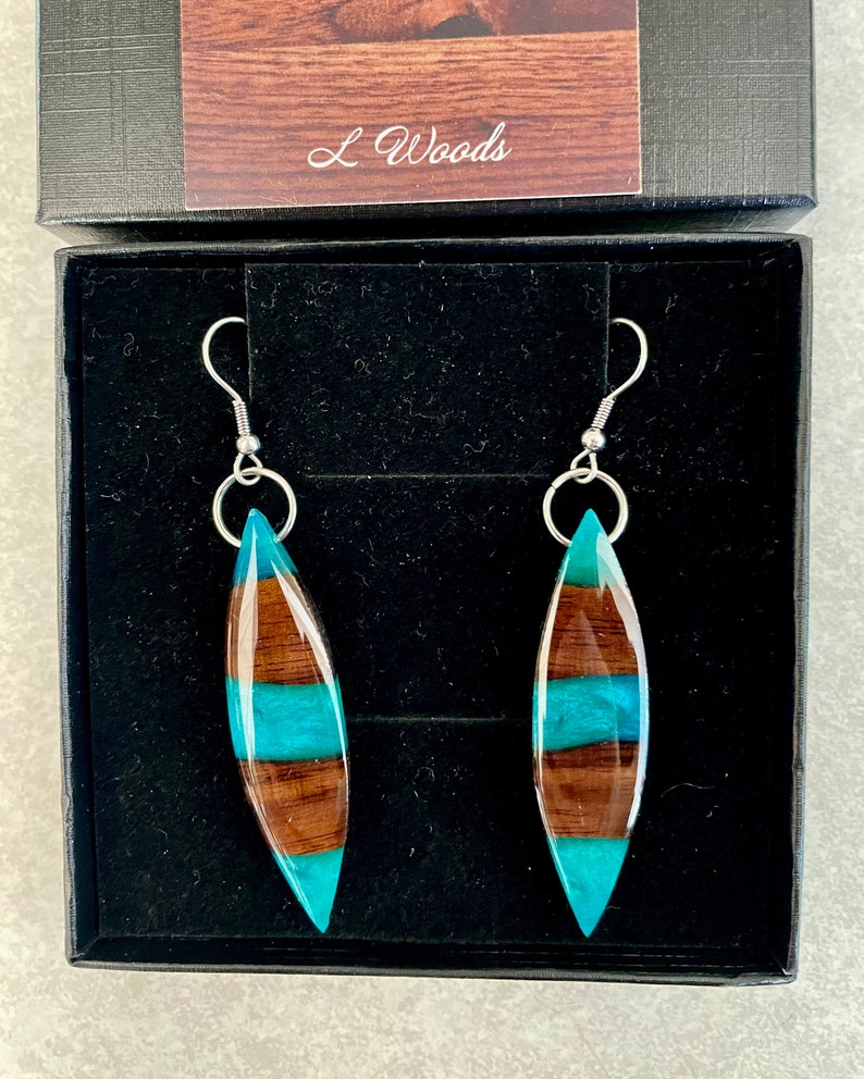 May include: A pair of teal and brown wood earrings with silver hooks. The earrings are in a black box with the text "L Woods" on the lid.