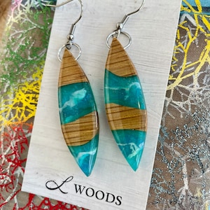 May include: A pair of teal and brown wood resin earrings with silver hooks. The earrings are shaped like teardrops and have a white swirl design inside the teal resin. The earrings are displayed on a white card with the text "L WOODS".