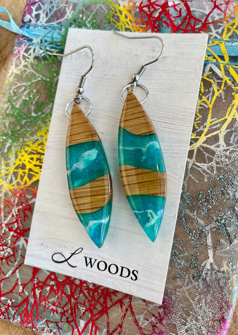 May include: A pair of teal and brown wood resin earrings with silver hooks. The earrings are shaped like teardrops and have a white swirl design inside the teal resin. The earrings are displayed on a white card with the text "L WOODS".