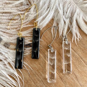 May include: Two pairs of dangle earrings with a rectangular resin charm featuring a dandelion seed head. One pair has a black resin charm and gold hooks, the other pair has a clear resin charm and silver hooks.