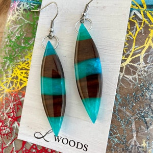 May include: A pair of teal and brown striped resin earrings with silver hooks. The earrings are shaped like a teardrop and have a glossy finish. The earrings are displayed on a white card with the text "L Woods" printed on it.