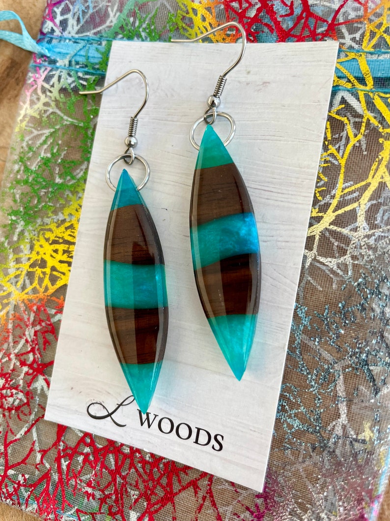 May include: A pair of teal and brown striped resin earrings with silver hooks. The earrings are shaped like a teardrop and have a glossy finish. The earrings are displayed on a white card with the text "L Woods" printed on it.