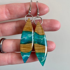 May include: A pair of dangle earrings with a wood and resin design. The resin is a teal blue color with white swirls and the wood is a light brown color. The earrings have silver hooks.