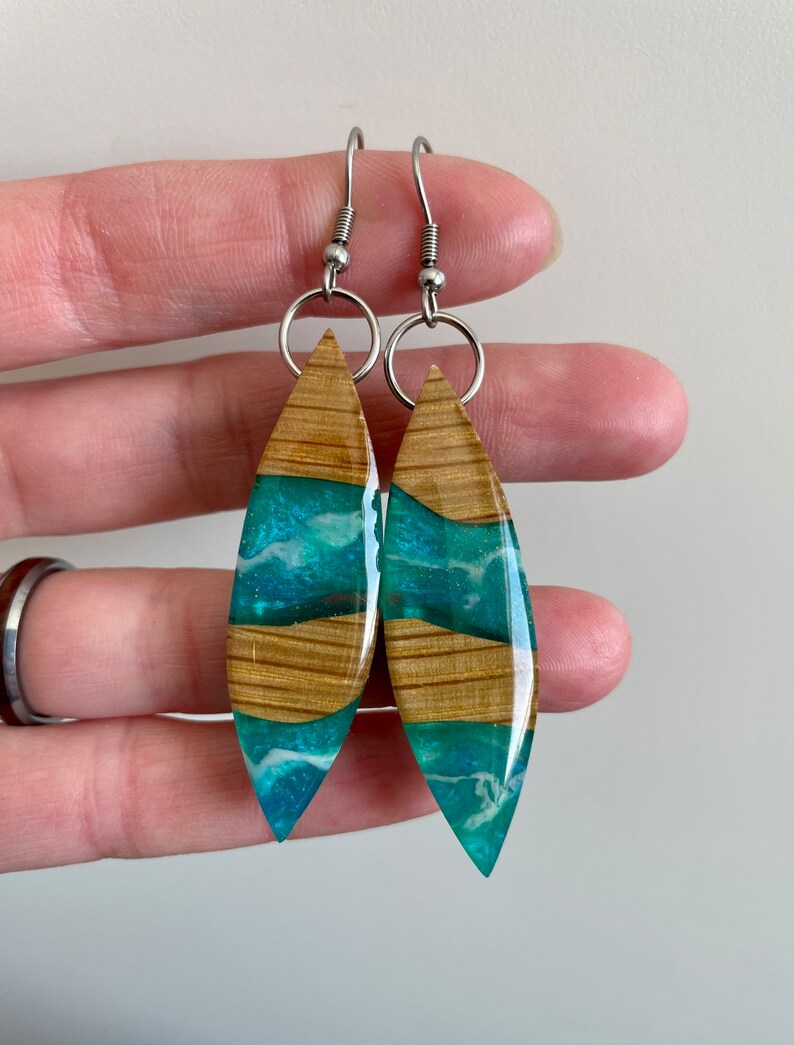 May include: A pair of dangle earrings with a wood and resin design. The resin is a teal blue color with white swirls and the wood is a light brown color. The earrings have silver hooks.