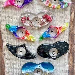 May include: Eight different bookmark designs featuring various colours, shapes, and patterns. Some bookmarks are heart-shaped, while others are crescent-shaped. Some bookmarks feature pressed flowers, while others feature a rainbow swirl pattern. All bookmarks have a hole in the centre for a finger to hold.