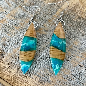 May include: A pair of teal and brown resin earrings with a wood grain pattern. The earrings are shaped like a teardrop and have silver hooks.
