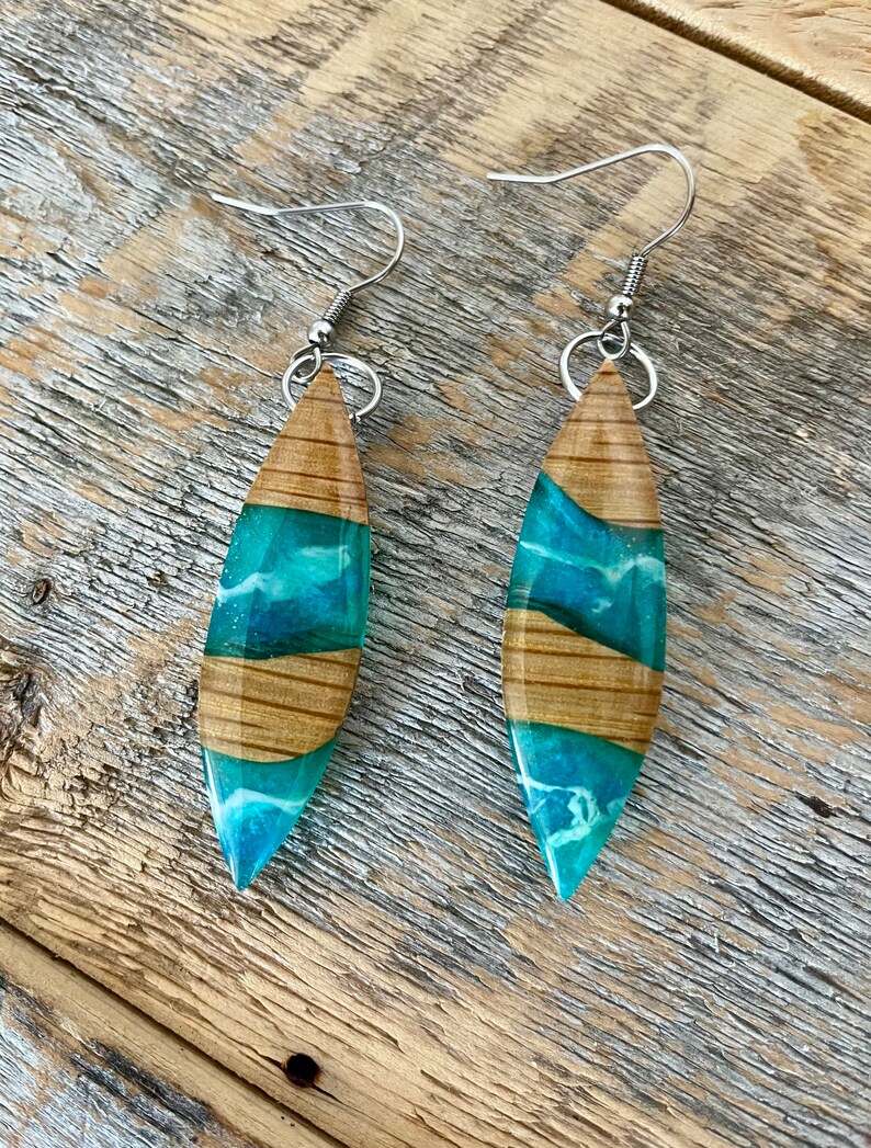 May include: A pair of teal and brown resin earrings with a wood grain pattern. The earrings are shaped like a teardrop and have silver hooks.