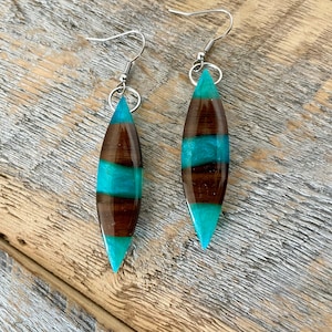 May include: A pair of dangle earrings with a teardrop-shaped resin pendant. The pendant is made with brown and teal resin and has a wood-like pattern. The earrings have silver hooks.