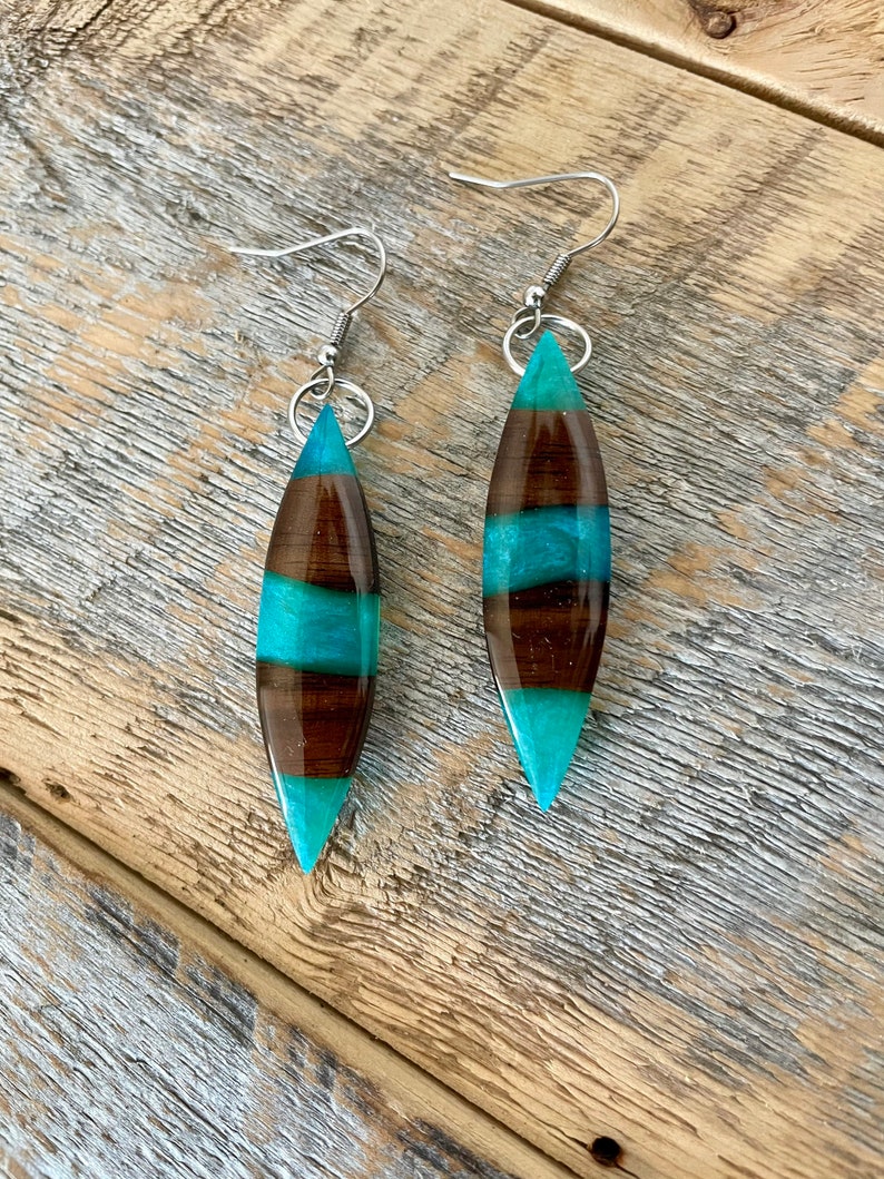 May include: A pair of dangle earrings with a teardrop-shaped resin pendant. The pendant is made with brown and teal resin and has a wood-like pattern. The earrings have silver hooks.