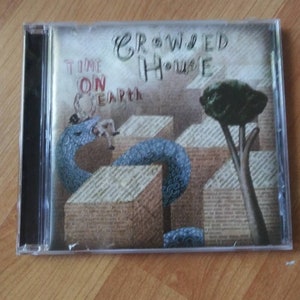 May include: A CD case with a cover featuring a blue snake wrapped around a building made of newspaper clippings. The text "Crowded House" and "Time On Earth" is written on the cover.
