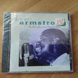 May include: A CD case with a black and white photo of Louis Armstrong singing into a microphone. The text on the case reads "Louis Armstrong The Best of the Decca Years Vol One the singer". A price tag is visible in the top right corner with the text "9.97 HMV $10.99".