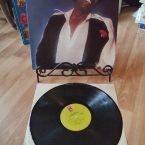 May include: A vinyl record album by Lou Rawls, titled "All Things in Time," with a portrait of Rawls on the cover. The record is displayed on a decorative stand, with a black vinyl record in the foreground.