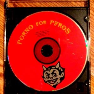 May include: A red CD with the text "PORNO FOR PYROS" in yellow letters. A cartoon devil with a red and black color scheme is in the center of the CD.