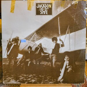 May include: A vintage vinyl record cover for the Jackson 5ive album titled Skywriter. The sepia-toned image features the five members of the group posing in front of a biplane. The album title and group name are printed in a black box at the top.