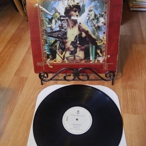 Pode incluir: Um álbum de vinil vermelho com uma colagem de imagens na capa. O álbum é intitulado "Disco's Greatest Hits" e apresenta um homem com uma coroa de espinhos na cabeça. O disco está em um tapete de toca-discos preto.